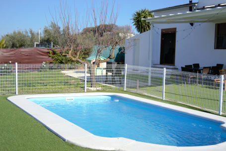 Piscina azul rodeada de césped y cercada, con una casa blanca de fondo.